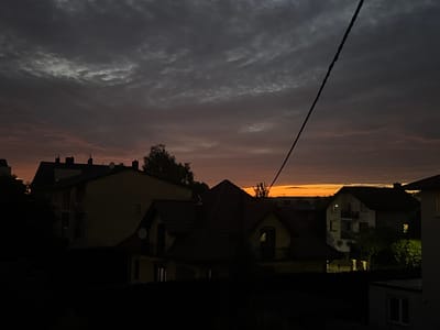 A twilight scene is depicted in Marki, Poland, with a dramatic sky transitioning from dark grey clouds above to hues of orange and pink near the horizon. Several houses, silhouetted against the fading light, are visible. In the foreground, a dark hedge obscures the lower portions of the buildings. The central focus is a house with a prominent, steep, red-tiled roof and dormer windows. To the left, a taller, lighter-colored building with multiple balconies stands. The background shows more residential structures and trees, all diminishing into darkness. A single black power line stretches diagonally across the upper right portion of the frame, cutting through the cloudy sky. The overall impression is a quiet, end-of-day urban landscape.