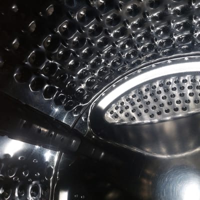 The image displays the interior of a domestic washing machine drum, constructed from polished, highly reflective stainless steel. The upper and left internal surfaces feature a textured pattern of numerous convex dimples. Towards the right, a curved segment of the drum wall is visible, perforated with a grid of small, circular holes. A dark gray or black plastic agitator paddle is positioned horizontally across the mid-left area, extending from the left edge towards the center. This paddle also exhibits small, circular perforations along its surface. Intense light reflections are evident on all metallic components, indicating a clean and illuminated interior. No individuals or foreign objects are present within the drum. The scene is located in Gimpo-si, South Korea.