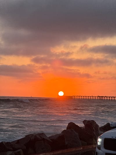 The image depicts a sunset scene at the Oceanside Pier in Oceanside, California, United States of America. The sun, a bright white disc, is setting over the Pacific Ocean, casting a vibrant orange glow across the sky and reflecting on the water. Dark, textured clouds are present in the upper portion of the sky, contrasting with the warmer tones below.

The Oceanside Pier, a long wooden structure with railings and lampposts, stretches horizontally across the middle ground of the image. Waves are breaking on the ocean's surface, creating white foam as they approach the shore. In the distance, a solitary sailboat is visible on the horizon.

In the foreground, dark, jagged rocks form a natural barrier. To the right, the front corner of a white vehicle is visible, with its headlights illuminated. The overall atmosphere is one of a serene and picturesque coastal sunset.
