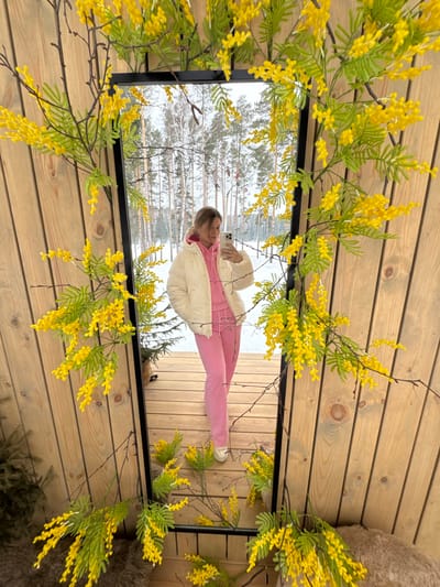 The image displays a full-length reflection of a female individual in a large, vertically oriented mirror, situated in Mishkino, Russia. The mirror features a black frame and is extensively decorated with artificial yellow mimosa-like flowers and green foliage, which also extends onto the light-toned wooden panel wall behind it. The reflected individual stands on a wooden deck or platform, wearing a cream-colored puffer jacket over a pink hooded sweatshirt and matching pink sweatpants, complemented by light-colored sneakers. She holds a light-colored smartphone with both hands, aimed towards the mirror, indicating the act of taking a selfie. The background reflected in the mirror shows a snow-covered forest composed of numerous tall, slender trees, under an overcast sky, consistent with a winter outdoor environment.
