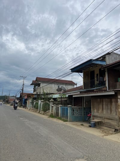 The image depicts a street scene in the city of Rantau Pauh, Indonesia, characterized by overcast skies and a row of buildings lining the right side of a paved road. Several motorcycles are visible on the road, with one rider in the foreground. The buildings appear to be residential or small commercial structures, exhibiting varied architectural styles and materials, including wood and concrete. Overhead, a complex network of power lines and utility poles crisscrosses the sky, suggesting an active electrical infrastructure. A decorative fence with blue and green accents separates the buildings from the road. The ground is a mixture of pavement and dirt. The overall impression is one of a functional, lived-in urban environment.