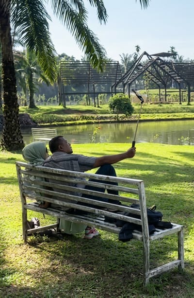 In Bintang Meriah, Indonesia, a couple is seated on a white slatted park bench, facing away from the camera. The man, wearing a checkered shirt and dark pants, is holding a selfie stick extended, with a smartphone attached at the end, capturing a photo of himself and the woman. The woman, wearing a light green hijab and a matching garment, is leaning against him. The bench is situated on a grassy area next to a body of water, possibly a pond or lake, which reflects the trees and the sky. In the background, there are skeletal, open-air structures that appear to be shelters or gazebos, surrounded by lush green foliage and palm trees. The overall scene depicts a relaxed moment in a park-like setting, with the couple engrossed in taking a photograph.