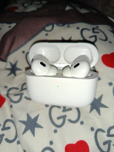 A white Apple AirPods Pro charging case is open, with two AirPods inside, resting on a patterned fabric. The fabric appears to have a repeating design of "GG" logos, stars, and red hearts. A small green LED light is visible on the front of the charging case, indicating its status. The image does not contain any information that would allow for the determination of its location in the city of Leninaul, Russia.