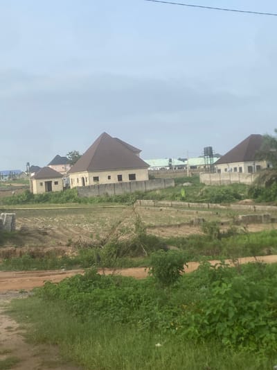 The image shows an outdoor scene in Iddo Tudunwada, Nigeria. In the middle ground, several buildings are visible, constructed with light-colored walls and brown, pitched roofs. One building, in particular, has a prominent pyramidal roof. These structures are partially enclosed by concrete block walls. In the foreground and surrounding the buildings, there is overgrown vegetation, including tall grasses and bushes. The ground appears to be a mixture of dirt and patches of barren land, with some areas exhibiting evidence of construction or development, such as stacked blocks and concrete foundations. In the background, more buildings and a water tower are discernible under an overcast sky. The overall impression is of a developing residential or commercial area.