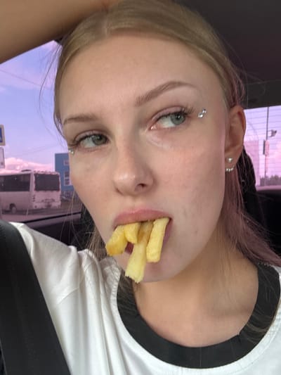 A close-up, eye-level medium shot captures a young woman with light blonde hair and green eyes as she holds several french fries in her mouth. She is looking to her right with a slight smirk. The woman is wearing a white t-shirt with black trim. She has small, sparkly accessories on her left eyebrow and a three-tiered earring in her right ear. The background shows a blurry view of a street scene, with a white bus and buildings visible. The sky is a soft purple and pink, suggesting either dawn or dusk. The overall impression is informal, likely taken inside a vehicle. The location is noted as Kondratovo, Russia.