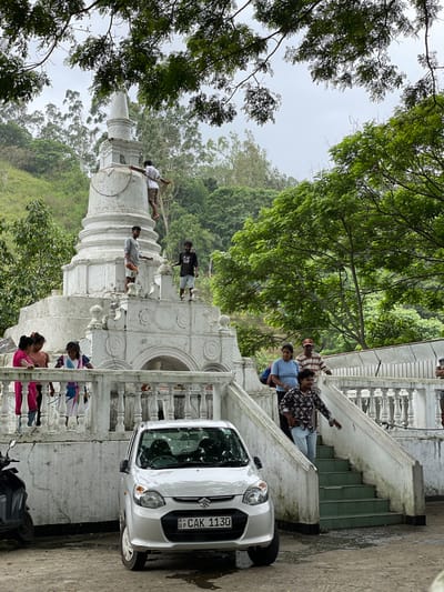 A group of people are gathered around a white stone structure in what appears to be Kumbalwela, Sri Lanka. The structure is tiered, with some people standing on its different levels. A white car, with the license plate "UP CAK 1130," is parked next to a flight of stairs near the structure. A black scooter is also present at the bottom of the stairs. The scene is framed by trees and foliage, with an overcast sky above.