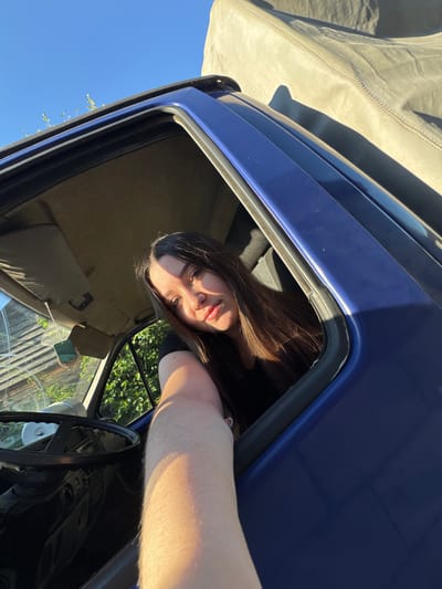 A young woman with long, dark hair is looking out of the passenger side window of a blue vehicle. She is wearing a black shirt. Her right arm is extended out of the window, casting a shadow on the exterior of the vehicle. The interior of the vehicle is visible, including the steering wheel and dashboard. Outside the vehicle, lush green foliage and a clear blue sky are visible. The vehicle appears to be parked outdoors, possibly in a residential area, given the presence of a fence-like structure in the background. The lighting suggests it is late afternoon or early evening, with the sun casting long shadows. The image was taken in the city of Krasnokamsk, Russia.