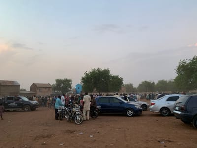 The image depicts a large gathering of people in Sokoto, Nigeria (13.049, 5.238). The scene appears to be an outdoor public event, possibly a political rally or a religious gathering. There are numerous parked cars and motorcycles, with several people standing around, seemingly waiting or observing something. A police truck is also present, indicating possible security measures. The environment is dusty and dry, with a few trees in the background. The sky is overcast with a pale blue hue, suggesting it might be early morning or late evening. The people are dressed in casual clothing, with some wearing traditional attire. There is no clear indication of specific actions or interactions, except for people standing and observing.