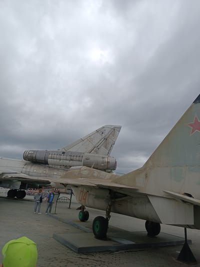 The image depicts an outdoor exhibition of aircraft under a cloudy sky. Two individuals, a man and a woman wearing casual attire, are observing the aircraft. The aircraft appear to be military jets, with one prominently featuring a red star insignia on its tail. In the foreground, a person wearing a bright green hat is visible from behind. The ground is paved with interlocking bricks, and the aircraft are displayed on elevated platforms or stands. The overall scene suggests a museum or memorial site dedicated to aviation history. The location is identified as the "Museum of military equipment UMMC" in Verkhnyaya Pyshma, Russia.