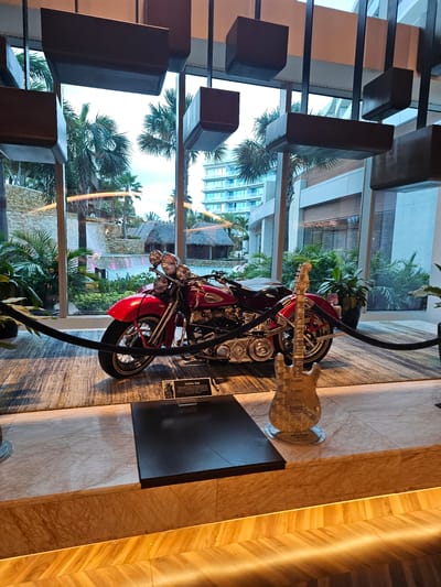 The image depicts an indoor display featuring a red motorcycle and a clear electric guitar. The backdrop is a large window offering a view of a tropical landscape, including palm trees and buildings, likely within a resort or entertainment complex. To the left, there's a decorative lighting fixture with rectangular elements. A sign reading 'Steve Vai' is visible next to the motorcycle. The scene is set at the Seminole Hard Rock Hotel & Casino in Hollywood, Seminole Reservation, Florida, United States of America.
