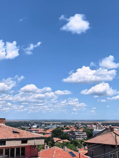 The image is a high-angle, full shot of a city skyline under a bright blue sky with scattered white clouds. In the foreground, several rooftops with traditional orange tiles are visible, some with solar panels. The middle ground shows a densely populated urban area with buildings of varying architectural styles, including apartment blocks and smaller houses, interspersed with trees. Beyond the city, a flat landscape stretches to the horizon, meeting the sky. The distant landscape appears to be a mix of agricultural fields and industrial structures, including silos. The scene suggests a sunny day in a city, possibly Svishtov, Bulgaria, given the presence of a dome on one of the buildings which could be a church. There are no people visible in the image.