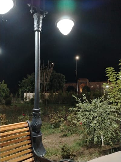 An outdoor nighttime scene in Khiva, Uzbekistan, features a street lamp in the foreground with a decorative base and a wooden slatted bench to its left. The lamp is illuminated, casting light on the surrounding area. Behind the lamp and bench, a park-like environment is visible with various trees and bushes, some of which appear to be rose bushes. In the mid-ground, a decorative metal structure resembling a basket or cage stands. Further in the background, a building with arched windows and a sign overhead is illuminated. Another street light is visible in the distance. The sky is dark, indicating night, with faint colored lights near the top of the frame. The ground appears to be a mix of dirt and grass.