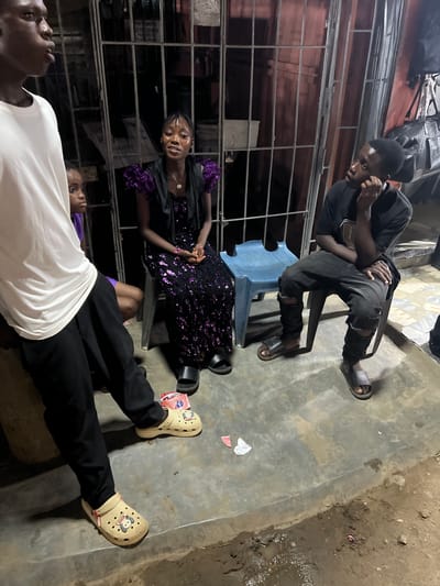 The image captures four individuals gathered in a dimly lit, semi-outdoor space in Ijegun, Nigeria. The ground consists of an uneven, stained concrete slab that transitions into a muddy, wet earth patch with small puddles in the foreground. Rusty metal grilles, possibly a gate or security barrier, form the background, revealing dark, indistinct walls and hanging bags behind them.

From left to right:
1.  A dark-skinned adult male is partially visible in the foreground left, wearing a white t-shirt and black trousers with light beige Crocs-style clogs on his feet. He faces right, with his mouth slightly open, appearing to speak or listen.
2.  Behind him, a dark-skinned child, likely female, is partially visible, wearing a purple top and looking towards the right.
3.  Seated on a dark chair in the center is a dark-skinned adult female, wearing a dark dress adorned with purple sequined or ruffled sleeves and a sequined skirt. Her hands are clasped, and she looks towards the left with a neutral expression. Dark slide sandals are on her feet. A light blue plastic stool is placed behind her to her right.
4.  On the far right, a dark-skinned adult male is seated on a dark chair. He wears a dark t-shirt and distressed dark jeans with rips at the knees, and dark slide sandals. His right hand is raised to his chin, and he looks upwards and to the left, indicating observation or contemplation.

Small pieces of white and red paper/debris are scattered on the concrete floor. The overall lighting is low, suggesting evening or night, with illumination from an unseen source.