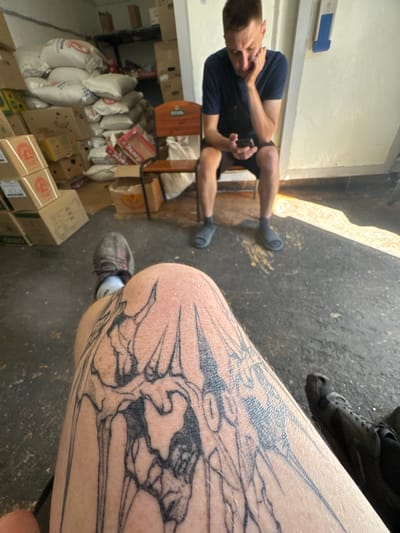 In Makiivka, Ukraine, a man with a dark blue t-shirt and dark shorts sits on a wooden chair, engrossed in his smartphone. Behind him, stacks of sacks and cardboard boxes fill a room, suggesting a storage or warehouse environment. In the foreground, a person's leg is visible, displaying a large tattoo of intricate, skeletal-like designs. The floor is a dark, rough surface, and sunlight streams in from the side, casting shadows. Another person's legs and feet, wearing dark socks and sandals, are also visible in the frame.