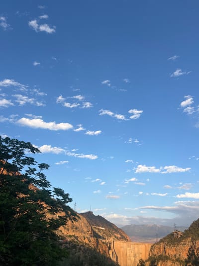 The image depicts a landscape in Bin El Ouidane, Morocco. A blue sky, speckled with scattered white clouds, occupies the upper portion of the frame. The right side features a rocky mountain range with visible electrical transmission towers and lines. A dam structure is built into the mountain. A tree with green foliage is visible at the bottom right corner. The environment appears to be a sunny, clear day.