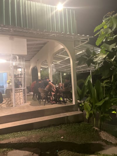 The image shows a group of young men sitting at tables under an open-air canopy, possibly a restaurant or cafe, at night. The scene is illuminated by artificial lights. In the foreground, there are stairs leading up to the dining area and a patch of grass. Behind the diners, there is lush green foliage. The architectural style suggests a modern, possibly tropical setting. The location is identified as Tabanan, Indonesia. The men are casually dressed and appear to be engaged in conversation or dining. One man is wearing a red and black checkered shirt, while others are in dark t-shirts. There is a glass enclosure to the left with what appears to be a bar or counter inside, featuring some equipment and flowers. Steam or smoke is visible in the air above the tables.