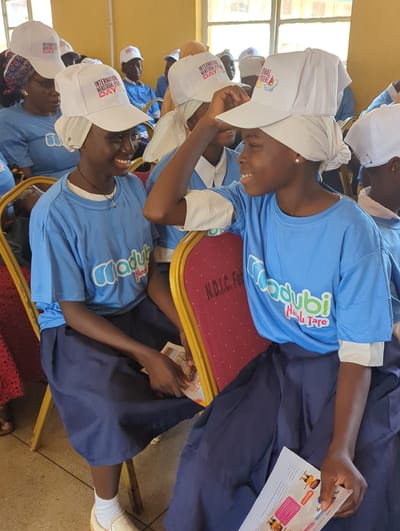 This media file captures a close-up, tilted view of several young Black individuals, likely students, wearing matching blue t-shirts and navy blue skirts. They are all wearing white caps with visible text and logos. The t-shirts have a colorful logo that reads "Dodybi Hana Tare". The caps appear to have text related to "International Menstrual Hygiene Day".

The subjects are seated, possibly on chairs with red cushioning and gold frames, suggesting an indoor setting. The lighting suggests it could be daytime, with natural light coming from windows visible in the background. The individuals are smiling and appear engaged, with one person in the foreground holding a piece of paper with text and images, possibly an informational flyer or schedule. Another person is adjusting their cap. The overall atmosphere seems positive and educational. Visible text on the caps and flyer indicates the event is related to International Menstrual Hygiene Day. The location context provided is Kakuri Gwari, Nigeria.