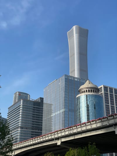 The image shows a low-angle view of modern architecture in Chaoyang District, China. The sky is a clear blue. Several high-rise buildings are visible. One is a curved, streamlined tower, another features a cylindrical glass structure. There are also rectangular glass buildings. Some greenery is present at the lower edge of the frame.
