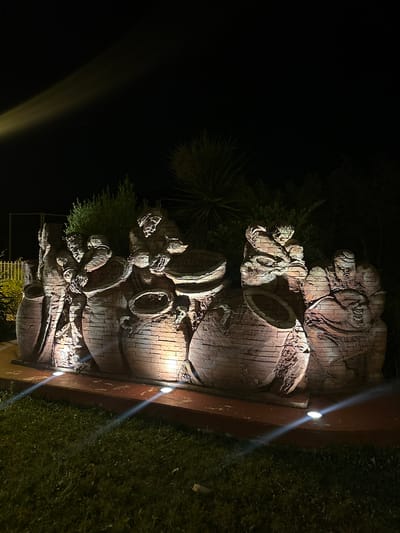 This is a nighttime photograph of a brick sculpture depicting several figures interacting with large clay pots. The sculpture is illuminated by several spotlights, creating dramatic shadows and highlights. The figures appear to be engaged in labor, with some holding tools or working with the pots. The sculpture is situated outdoors, with a grassy area in the foreground and lush vegetation behind it. The overall impression is one of a rustic and artisanal scene. This sculpture is located in Anduze, France, likely a public art installation or part of a garden.
