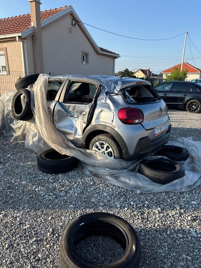 In the city of Mojanovići, Montenegro, a silver Citroën C3 is parked on a gravel lot. The car has sustained significant damage, with its rear end severely crumpled and the rear windshield shattered. The vehicle is partially covered in a translucent plastic sheeting. Several tires are scattered around the car, with two placed in front and two resting on the damaged rear section. In the background, a light-colored house with a red tile roof and a black Audi A4 are visible. The scene appears to be outdoors, under a clear blue sky, likely in a residential or semi-industrial area, given the presence of houses and a fence.