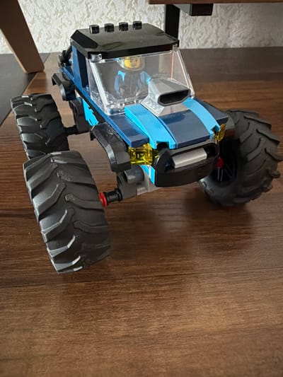 A toy monster truck, primarily blue and black with clear plastic windows, is positioned on a wooden surface. The truck has large, knobby tires. The scene appears to be indoors, possibly on a table or floor with a wood grain pattern. There are no people visible. The lighting suggests it could be daytime, but no specific time of day can be determined. No weather conditions are apparent. The location context is Yablonovskiy, Russia, but this is not visually evident in the image. There is a small yellow label with black text on the truck, but the text is too small and blurry to be read.