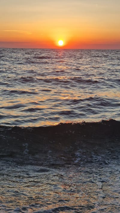 This media file captures a serene sunset over the Adriatic Sea in Zadar, Croatia. The sun, a vibrant orb of orange and yellow, hovers low on the horizon, casting a warm glow across the sky and reflecting on the water's surface. The sea itself is characterized by gentle, undulating waves, their crests catching the last rays of sunlight, creating a shimmering, almost textured appearance. The scene is divided vertically, with the left side dominated by the soft hues of the sunset sky and the sun, and the right side by the rich, darker tones of the rippling water. The overall impression is one of tranquility and natural beauty. The time of day is clearly sunset, with the low angle of the sun and the warm, diffused light. No people or specific objects are visible, focusing the attention entirely on the natural elements of the seascape. The location is suggested to be Zadar, Croatia, likely a coastal area. There is no visible text in the image.