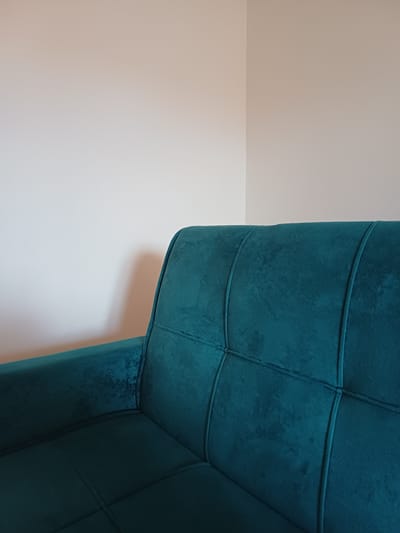 This image shows a close-up, abstract view of a teal tufted sofa positioned against a white wall. The sofa has a velvet-like texture with distinct stitched patterns forming squares across its surface. The lighting creates shadows and highlights on the tufted sections, emphasizing their three-dimensional quality. The white wall provides a minimalist backdrop, with a subtle shadow cast by the sofa suggesting it is near a light source. The perspective is angled, with the sofa dominating the right side of the frame and a large expanse of white wall on the left. No people or specific activities are visible. The image appears to be taken indoors, in a well-lit room. There is no visible text in the image. The overall impression is one of modern interior design.