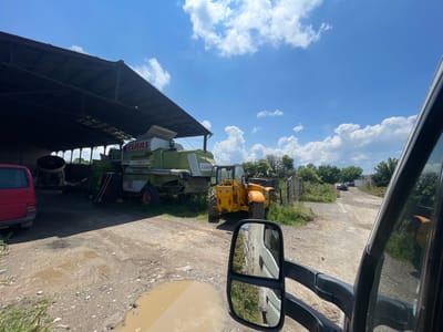 The image presents a daytime scene at what appears to be a farmyard or industrial storage area in Gorni Bogrov, Bulgaria. The perspective is from inside a vehicle, likely a van or truck, with a large black side mirror and part of the window frame occupying the right side of the frame.
In the midground, under a large, open-sided shed with a dark roof, a green combine harvester is prominently parked. Visible text on the combine includes "CLAAS", "MAXI", "108", and "SE". To its left, a red van is partially visible. Next to the combine harvester, a yellow telehandler is parked on a dirt and gravel surface, near a wooden fence and some overgrown grass.
The ground is unpaved, showing tire tracks and several muddy puddles that reflect the bright sky. Beyond the machinery and fence, a dirt track or road stretches into the background, flanked by lush green trees and bushes. A dark-colored car can be seen further down this track.
The sky is a vibrant blue with scattered white, cumulus clouds, indicating a bright and sunny day. No people are visible, and the scene depicts parked vehicles and machinery rather than active work.