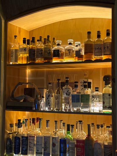 A close-up shot depicts a backlit bar display showcasing a variety of alcoholic beverages. The left shelf is lined with Japanese whiskey bottles, including Suntory Toki and Yamazaki. The central shelf features clear liquor bottles, some identified as vodka, and a small "Fitos" glass, in addition to a decorative shark jaw and books. The right shelf is stocked with tequila and mezcal bottles, including labels such as "Tritero", "Liberador", "Maachee", and "G4". The image was likely taken in a bar in Oaxaca City, Mexico, given the types of alcoholic beverages prominently displayed.