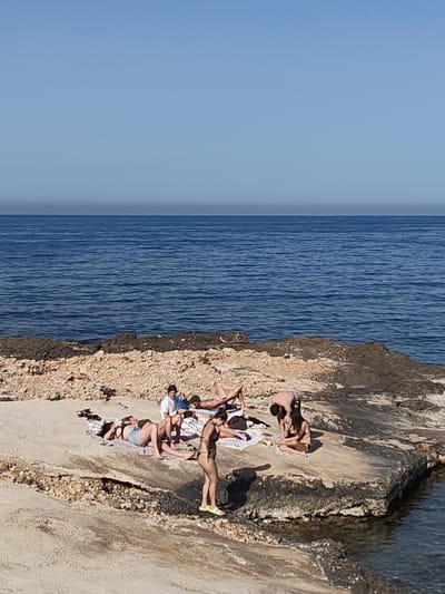 The image depicts a group of people on a rocky outcrop at a beach in Rotes, Spain. The sky is clear and blue, and the sea is a deep blue color. Several people are sunbathing on towels. A person in a white shirt is sitting amongst the sunbathers. Another person is bending over, while another person is seated. One person is standing on the rocks wearing a dark bikini and yellow sandals.