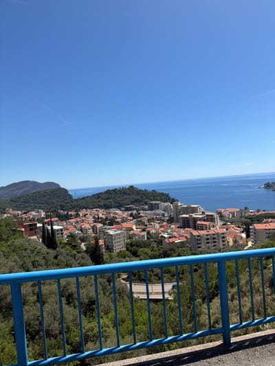 The image presents a high-angle view of a coastal town nestled between lush green hills and the sparkling blue Adriatic Sea. In the foreground, a vibrant blue metal railing runs horizontally across the frame, with vertical bars spaced evenly. Beyond the railing, the town unfolds with a dense cluster of buildings characterized by orange tiled roofs, interspersed with modern apartment blocks and some older, unfinished structures. The buildings are surrounded by a thick canopy of green trees, with olive trees and cypress trees being prominent.
Further into the scene, a winding road can be seen cutting through the greenery, with a single blue car driving on it. The landscape rises into rolling hills covered in dense, dark green vegetation. In the distance, the vast expanse of the Adriatic Sea stretches to the horizon, meeting a clear, bright blue sky. A faint white contrail is visible in the sky, indicating the presence of an aircraft. The overall impression is one of a sunny day in a Mediterranean coastal area.
While the image depicts a generic coastal town, its features are consistent with the landscape and architecture found in the region of Stupovi, Montenegro, which is known for its scenic coastal beauty, hilly terrain, and proximity to the sea.