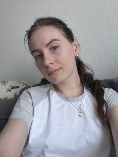 An indoor close-up image taken in Yanino-1, Russia, features a young adult female centered in the frame, captured from the chest up. She has dark brown hair, styled with visible gray strands near the forehead and temples, pulled back into a ponytail on her right side. Her complexion is light, and she has light-colored eyes with faint undereye discoloration. A small brown mole is visible on the left side of her neck. She wears a white t-shirt with light grey raglan sleeves, a gold-colored hoop earring in her visible right ear, and a thin gold-colored chain necklace with a circular pendant containing a small clear stone. The subject is looking directly at the camera. The background consists of a light grey, textured wall. To the subject's left, a light-colored pillow patterned with cartoon corgi dogs rests on a grey upholstered surface.