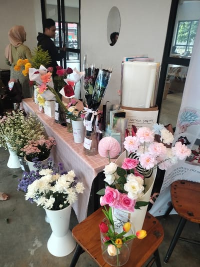 This image captures a vibrant indoor flower stall or market display, likely in Dramaga, Indonesia. The setting appears to be a brightly lit commercial space, possibly a cafe or a pop-up market, with a concrete floor and white walls.
In the background, two people are visible near a glass door or window. One person wears a light brown hijab and a green top, while another, a male in a black hoodie, is reflected in a small oval mirror mounted on the wall. Their backs are mostly to the camera, and their emotions are not discernible.
The central focus is a long table draped with a light pink and white checkered tablecloth, laden with a diverse array of fresh and artificial flowers. Bouquets of roses in pink, white, and red are prominent, alongside arrangements of white and purple chrysanthemums, baby's breath, and other floral varieties. Some bouquets are elegantly wrapped in white or black paper, the latter featuring "LOVE YOU" text, while others are presented in vases, bottles, and white buckets on the table and floor.
Notable objects on the table include small bottles, a brown paper bag labeled "sac en papier" (French for "paper bag"), and various other decorative elements. To the right, two wooden stools are placed, with one holding a clear glass vase containing artificial red and yellow tulips. A white standee or banner is partially visible, displaying text like "FLOWERS BOUQUET FRESH," "PREORDER," and "15.000," likely indicating pricing in Indonesian Rupiah, along with contact information such as "whatsapp."
The scene suggests an event or daily operation of a florist, showcasing a wide selection of flowers for sale. The bright lighting indicates it is daytime.