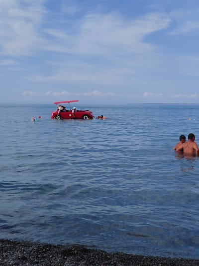 The image depicts a scenic view of a body of water, likely a lake, with a clear blue sky and scattered white clouds. The setting appears to be a beach with dark pebbles on the right side of the frame. Two men are visible in the water, immersed up to their chests, with their backs to the camera. In the middle ground, a bright red novelty boat shaped like a car is in the water, with several people on board. One person appears to be steering, and others are seated. The boat has yellow wheels. A colorful buoy is also floating in the water near the boat. The overall impression is a recreational scene on a sunny day. The lighting suggests it is daytime, likely midday or afternoon, given the bright sky. The location is identified as Akshi, Kazakhstan.