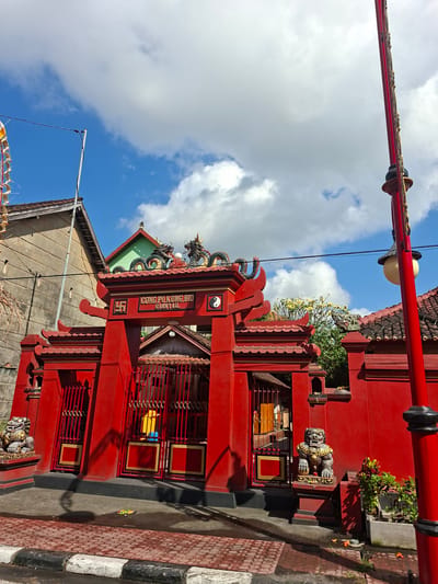 The image shows the bright red entrance to a temple in Gianyar, Indonesia. The temple's gate is ornately decorated with traditional Chinese architectural elements, including curved eaves and dragon statues on the roof. The temple's name, "CONG PO KUNG BIO GIANTAR," is visible above the entrance. Flanking the entrance are two stone lion statues, commonly found in front of temples and palaces. The street in front of the temple is paved with red bricks, and there are potted plants and fallen flowers on the ground. The sky above is a vibrant blue with scattered white clouds, suggesting a sunny day. A red lamppost stands to the right of the temple entrance. The overall scene conveys a sense of vibrant cultural and religious heritage.