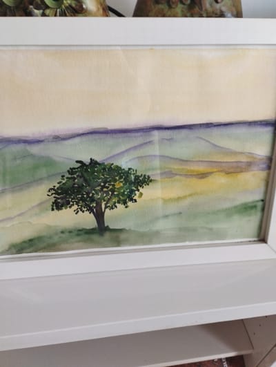 This is a watercolor painting in a white frame, depicting a serene landscape. The scene is a rolling countryside with layered hills painted in soft shades of green, yellow, and purple. In the foreground, a single, dark green tree stands prominently on a grassy knoll. The sky is a pale yellow, suggesting either early morning or late afternoon light. The overall mood of the painting is peaceful and tranquil. There is no visible text in the image. The location context of Pardes Hana-Karkur, Israel, is not visually indicated within the painting itself.