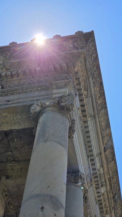 The image shows a low-angle view of the corner of an ancient stone structure, likely a temple, with ornate carvings and columns. The sun is shining brightly from the upper left, creating lens flare and highlighting the clear blue sky. The stonework appears weathered and is decorated with intricate patterns, including floral motifs and geometric designs. Two large, cylindrical columns are visible, supporting an architectural element with decorative capitals. The structure is bathed in sunlight, with shadows indicating the time of day and the sun's position. The overall impression is one of historical grandeur and architectural detail, set against a bright, clear sky. The location is the Temple of Garni, a classical temple in Garni, Armenia, which is a Greco-Roman colonnaded monument.