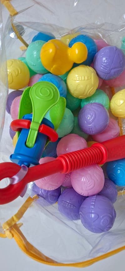 The image shows a close-up of a clear plastic bag filled with colorful plastic balls and two toy implements. The balls are various shades of pink, yellow, purple, and blue, and some have textured patterns. A yellow duck-shaped toy is visible among the balls. There is also a green scoop-like toy with red and blue plastic parts, and a red plastic toy with a coiled handle. The bag has yellow trim and appears to be a product meant for children. The background is a plain white surface. No specific details about the location in Orimattila, Finland, are provided or discernible from the image.