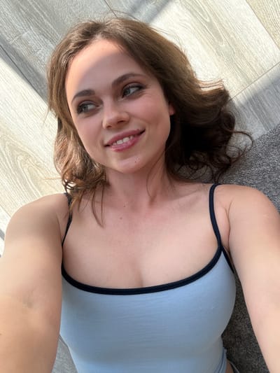 A woman is lying on her back in an indoor setting. She has fair skin, brown hair, and is wearing a light blue tank top with black trim. Her face is visible, and she is smiling. The background includes light-colored wooden flooring and a gray carpet or rug. The location cannot be determined as Balatonlelle, Hungary.