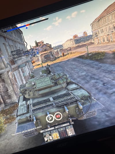 This is a screenshot from a video game, likely War Thunder, depicting a military tank in a city environment. The tank is green and appears to be in a damaged state, as indicated by the "The vehicle will be repaired in 00:02" text on the bottom left of the screen. The surrounding environment is urban, with buildings that appear somewhat dilapidated. There are no people clearly visible in the image. The game's user interface is also present, showing player information, ammunition counts, and a reticle. The scene is set during daylight with a clear sky. The mention of Kyustendil, Bulgaria, is not verifiable from the image content itself, as this is a virtual representation within a game.
