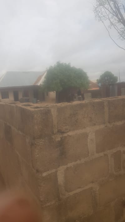 The image shows a building with a light-colored roof and a dark-colored wall in the foreground. The building appears to be a house or a school. There are trees and other vegetation around the building. The sky is overcast. The image is taken from a slightly elevated angle, looking down at the building. The foreground is dominated by a rough, light brown brick wall. In the background, a building with a light grey corrugated metal roof stretches horizontally. To the right of the building, there is a large, leafy green tree. Further to the right, another tree is visible, and beyond that, a corrugated metal fence. The overall impression is of a residential or semi-rural area, possibly in the town of Wukari, Nigeria.