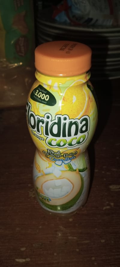 The media file captures a single, yellow and green plastic bottle of Floridina Coco beverage, standing upright on a dark, possibly wooden, surface. The bottle has a ribbed orange cap, which bears an expiry date (partially visible as "13-05 YEAX").
The label on the bottle is vibrant, featuring illustrations of sliced oranges and a cracked coconut revealing its white flesh, along with ice cubes, indicating the drink's flavor profile of orange and coconut. Key text on the label identifies the product as "Floridina Orange Coco" and highlights its contents: "Real Orange Pulp! & Coco Bitz." Below the main product name, "VIE C" is also visible, likely referring to Vitamin C.
A prominent green patch on the upper part of the label displays pricing information: "Harga Retail Rp 3.000 botol (Pulau Jawa)," which translates to a retail price of 3,000 Indonesian Rupiah per bottle, specifically for Java Island, Indonesia.
The background is out of focus, suggesting an indoor setting with artificial lighting, possibly indicating evening or nighttime. To the left, a blurry green and yellow package, potentially a snack bag, is visible. To the right, stacked dark objects, possibly plates or bowls, are partially seen.
There are no people or discernible activities depicted. The image is a static shot of the product. The visible text clearly indicates the product's market as Java Island, Indonesia, and offers no specific visual cues or details related to Aral, Kyrgyzstan, the provided location context.