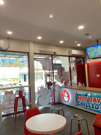 The image shows the interior of a store in Kecamatan Piyungan, Indonesia, located at coordinates -7.837, 110.474. The store is brightly lit with a white ceiling containing several recessed lights. The walls are painted white and red, and the floor is tiled. There are large glass windows and doors that open to an exterior view of a residential area. The store features several cartoon images of snowmen and a woman, all wearing crowns. The front counter is decorated with a large sign that reads "Hot Day Chilled." There are three white plastic chairs and two silver metal stools situated around a white round table. A red barstool is next to the window. A black and white television is hanging on the red wall, showcasing a promotional image of a dessert. The store is empty and unoccupied.