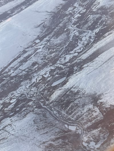 An aerial view depicts a landscape of striated rock formations with a network of waterways. Light-colored channels meander through dark, textured rock, creating a contrast between the reflective water surfaces and the solid ground. A straight, narrow path intersects the waterways. A small lake, or a widened section of a channel, stands out with its larger, smoother surface. The overall composition suggests a rugged, geological area shaped by erosion and water flow.