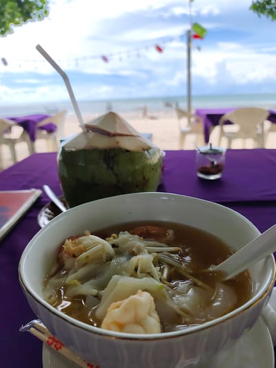 A close-up shot focuses on a bowl of food and a coconut drink on a table at the beach. The bowl, in the foreground, contains a clear broth with dumplings, noodles, and shrimp. A white spoon rests in the bowl. To the left, a halved coconut with a straw is placed on the table. The table is covered with a purple cloth.
In the background, chairs and tables are set up on a sandy beach, with the ocean and a cloudy sky visible. The scene suggests a relaxed dining experience by the sea, possibly during daytime. The overall mood is tranquil and inviting, highlighting a tropical setting in Sakhu, Thailand. No visible text is present in the image.