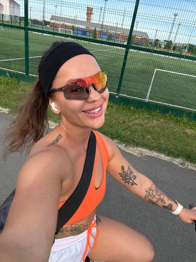 A young woman with brown hair pulled back into a ponytail and wearing a black headband is cycling outdoors in Yuzhnouralsk, Russia. She is wearing orange sports bra, white shorts with orange drawstring, and white wireless earbuds. She has sunglasses with reflective lenses and tattoos on her left shoulder and arm. The background shows a fenced-in sports field with a soccer field and buildings in the distance. The lighting suggests it is late afternoon or early evening.