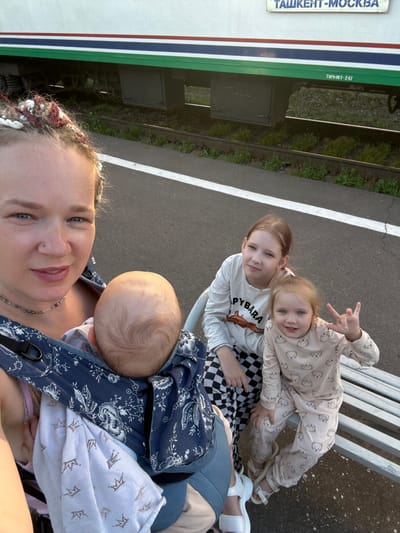 A woman is taking a selfie at a train station in Uzunovo, Russia. She is wearing a patterned baby carrier with a baby inside. To her right are two girls sitting on a bench. The older girl is wearing a white shirt with "PYBARA" printed on it, and checkered pants. The younger girl is wearing a pajama set with teddy bear prints. The train is visible in the background with "ТАШКЕНТ-МОСКВА" written on it.