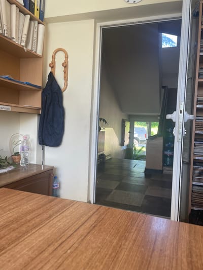 The image depicts an interior view of an office or reception area, likely located in the city of Elena, Bulgaria. In the foreground, a portion of a wooden desk is visible. To the left, a shelf unit holds various binders and documents. A dark blue jacket is hanging on a wooden coat rack mounted on the wall. Below the shelves, a wooden cabinet is present, with a bottle of water and a plant on its surface. The doorway in the center of the image reveals a staircase leading upwards and an exit with natural light and greenery visible outside. A light-colored vehicle is parked outside the exit. To the right of the doorway, a tall shelf unit is filled with books or files, with some labels visible. The floor inside the office appears to be tiled in a checkered pattern.