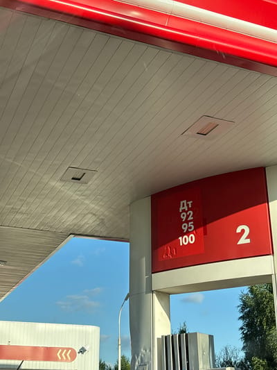 This is a photograph of a gas station canopy in Kirovo-Chepetsk, Russia, on a clear day. The canopy has a red trim and a white corrugated ceiling. A red sign displays fuel prices: "ДТ" (diesel), "92", "95", and "100". To the right of the fuel prices, the number "2" is prominently displayed. The sun is casting shadows on the canopy, indicating it is likely daytime. In the background, a blue sky with scattered clouds is visible, along with parts of a white building and trees. There are no people or vehicles clearly visible in the frame. The image appears to be taken from a low angle, looking upwards at the gas station infrastructure.
