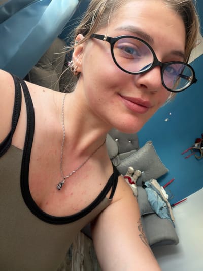 A woman with blonde hair and glasses is taking a selfie in an indoor setting. She is wearing a tank top and a necklace. The background is a blue wall with some furniture and objects. The image appears to be taken in a residential area. There is no information about the location being in Большая Шильна, Russia.